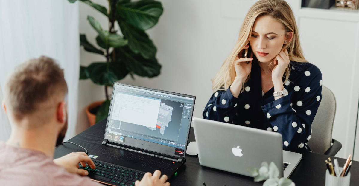 Two coworkers using laptops and phones while working together in a modern office setting.