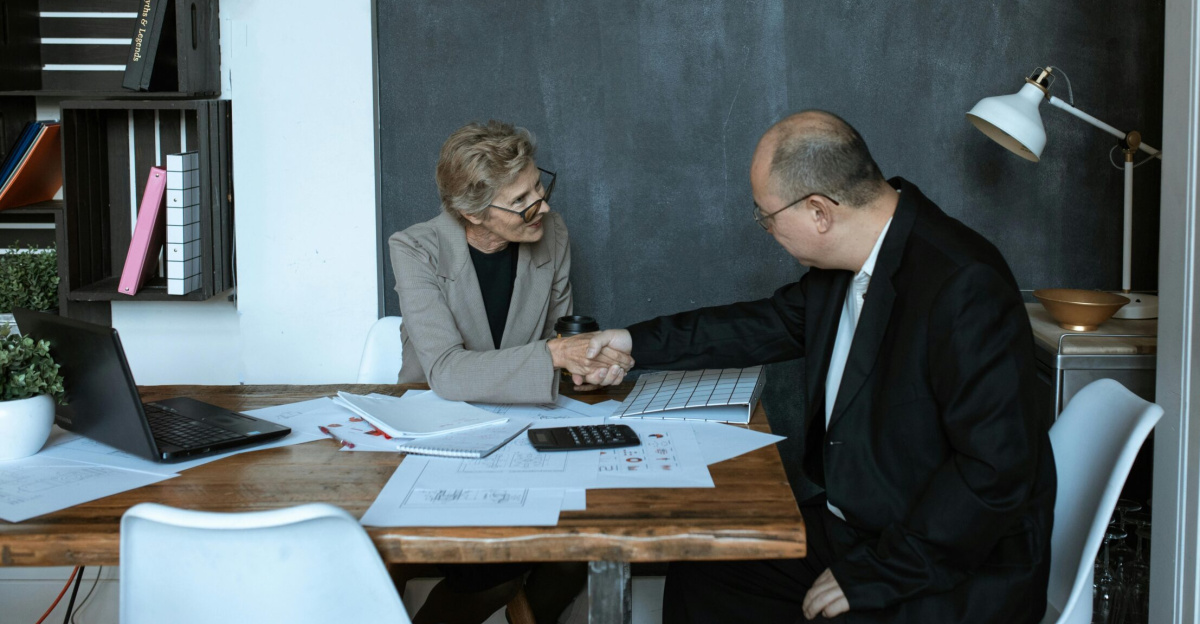 Two professionals engaged in a handshake across a desk in a modern office setting.
