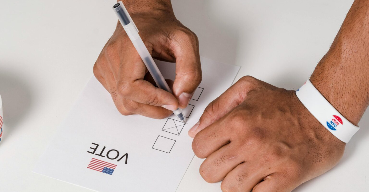 A person wearing a wristband fills out a voting ballot with a pen. Voting stickers visible.