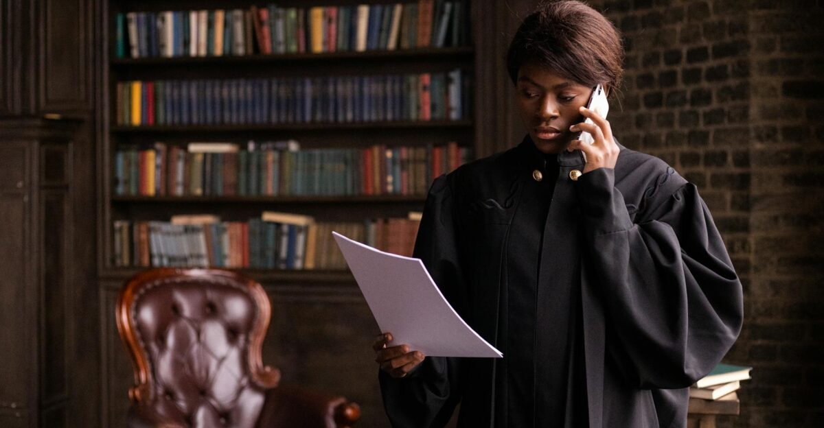 African American judge in traditional robes holds a document and talks on the phone in a law library