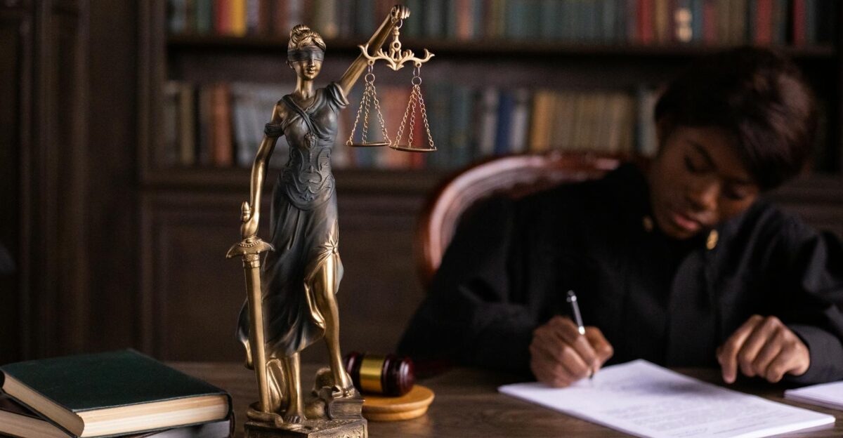 A focused judge writing on documents beside a Lady Justice statue in an office