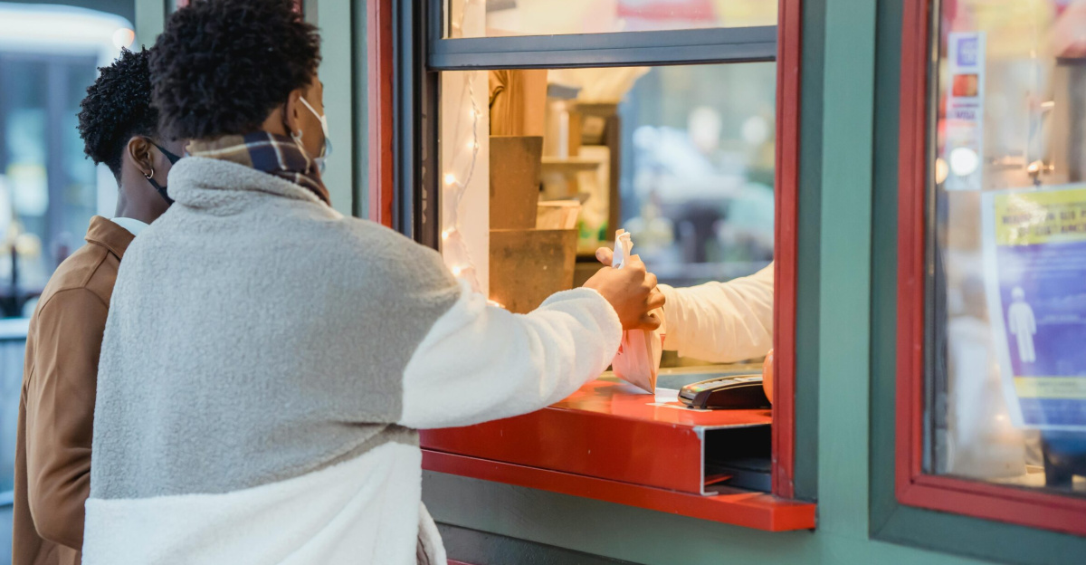 Side view of anonymous African American male couple in medical masks buying food on street while standing near window of store