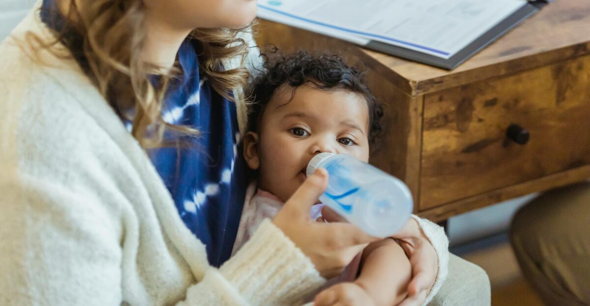 Unrecognizable mother bottle feeding cute little African American baby in hands while sitting near wooden drawer in light room at home