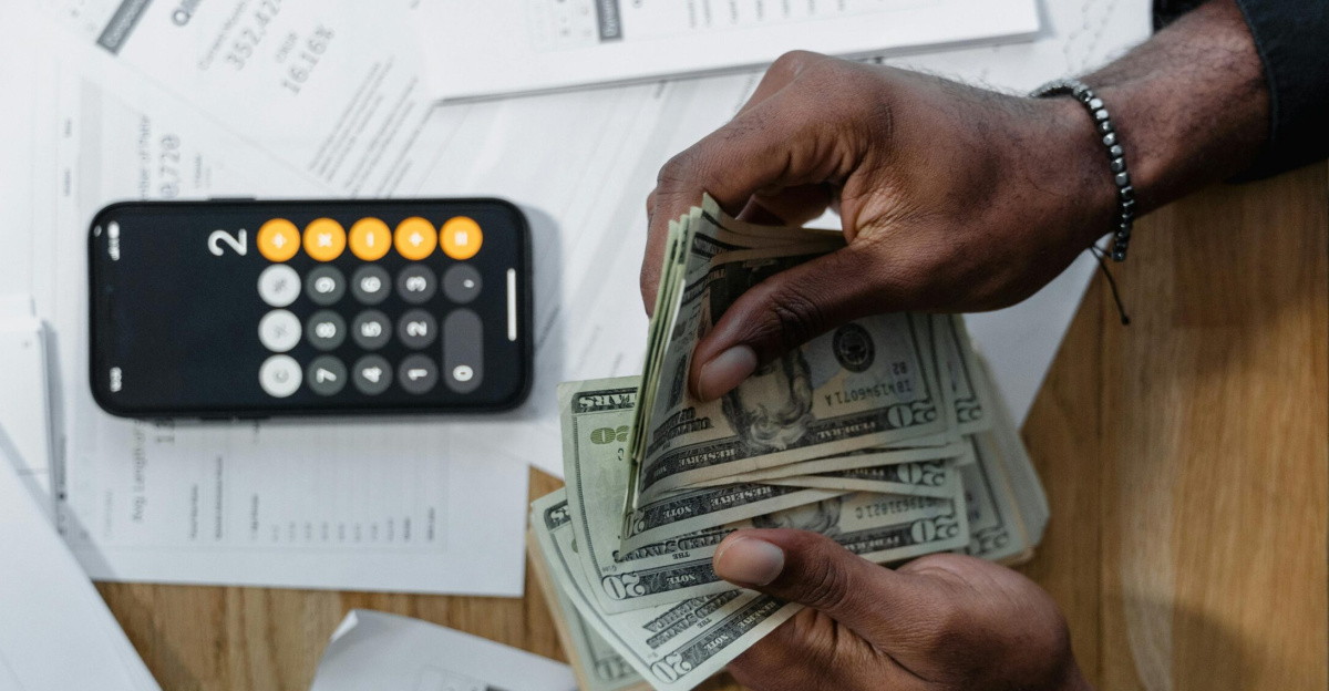 Person counting dollar bills over documents with a smartphone calculator on the desk.