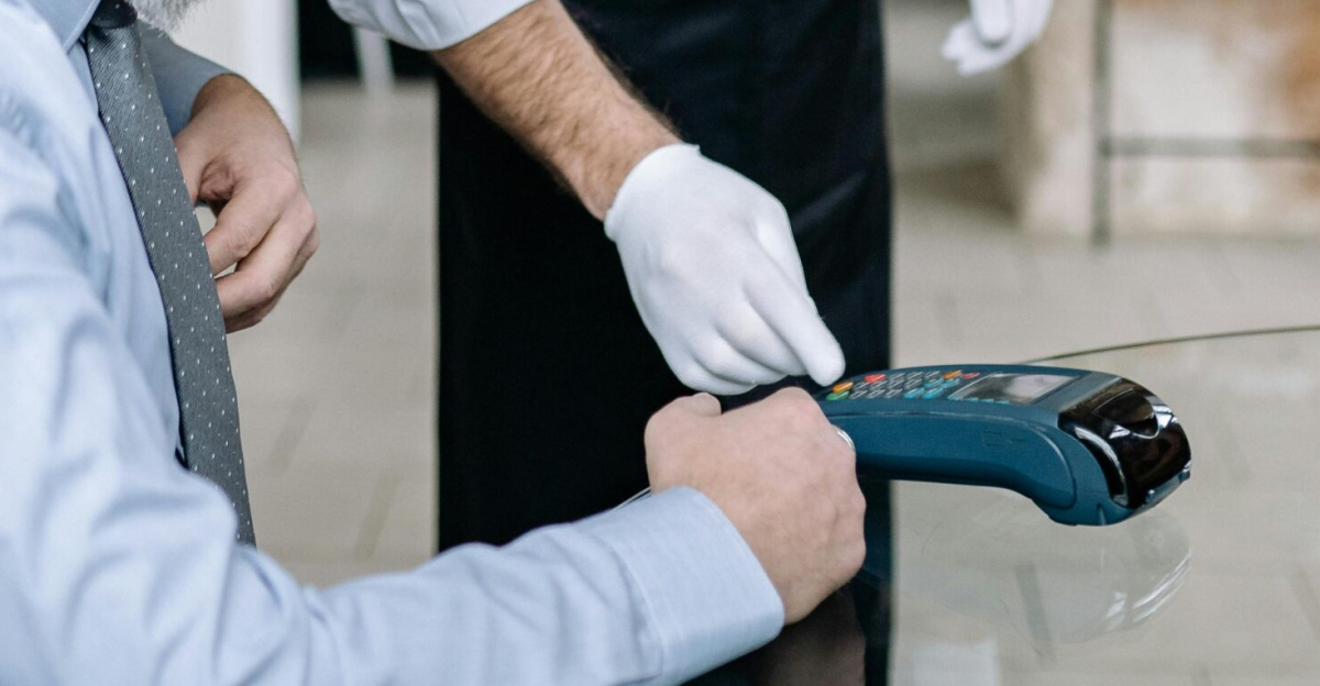 A customer pays using a card reader in a restaurant. The waiter wears a mask and gloves.