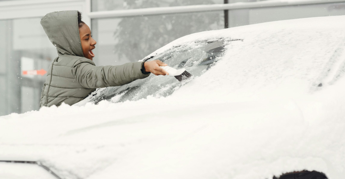 A woman in a gray coat clears snow from a car windshield on a cold winter day.