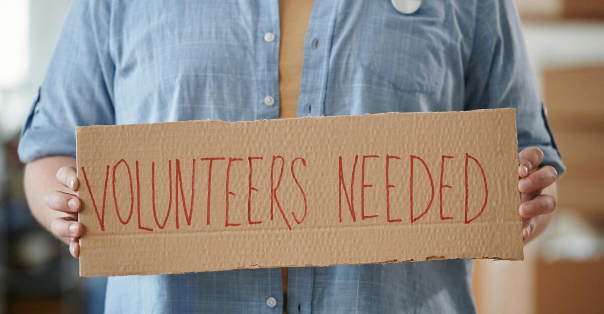 Person holding a sign reading Volunteers Needed to encourage community support