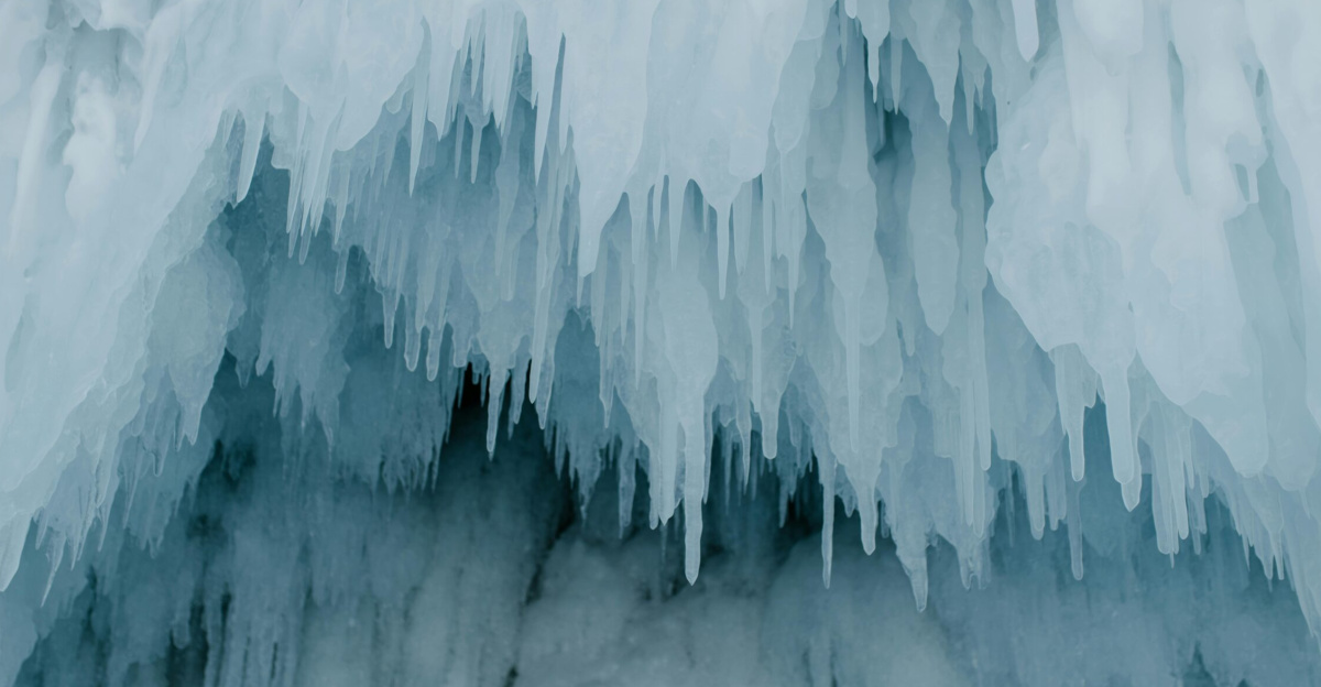 A dramatic close-up of sharp icicles hanging at a cave entrance in a winter setting.