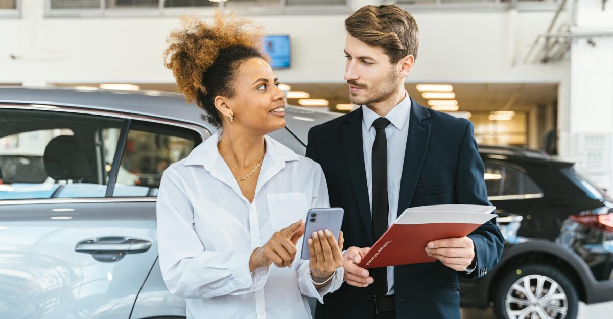 African American woman and Caucasian man discuss car purchase at dealership using smartphone