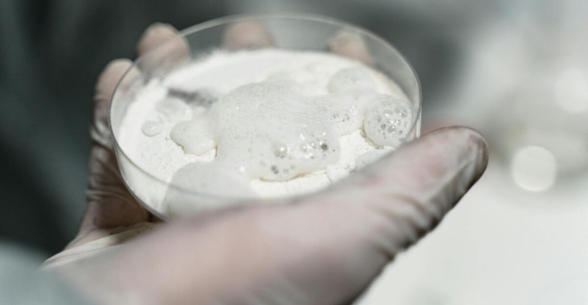 Close-up of a gloved hand in a lab holding a petri dish with a foamy substance possibly for scientific analysis