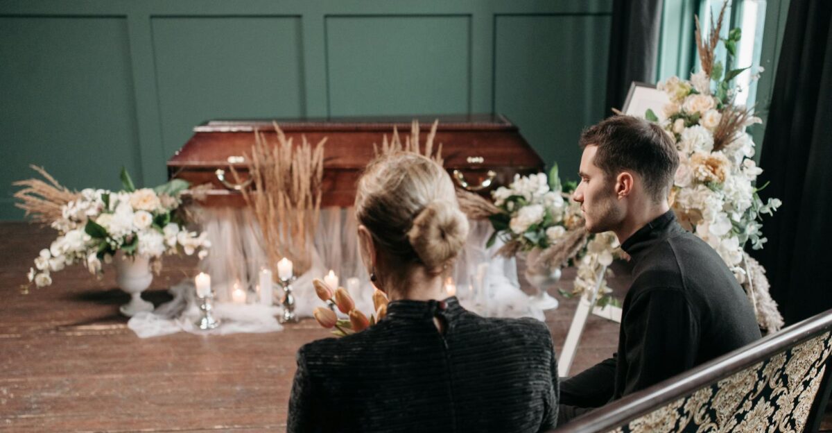 A funeral service with a wooden coffin adorned with flowers and candles attended by two mourners
