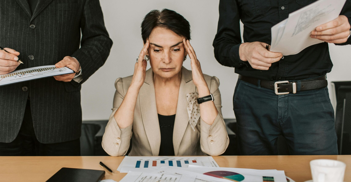 A stressed businesswoman holding her head with colleagues holding documents in a corporate office setting.