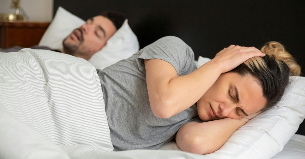 A woman looks frustrated in bed as her partner snores loudly, highlighting sleep disturbance issues.