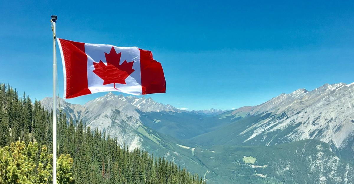 Canadian flag waving atop a scenic mountain view with blue skies and lush greenery.