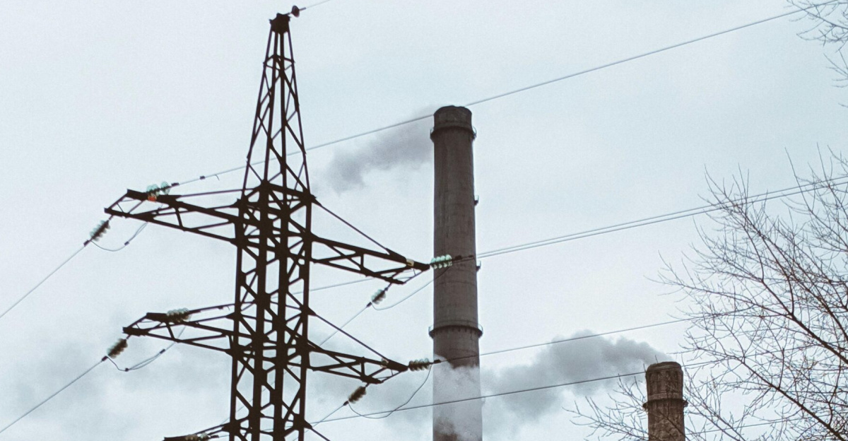 A power plant with towering smokestacks emitting smoke beside a high voltage pylon on an overcast day.