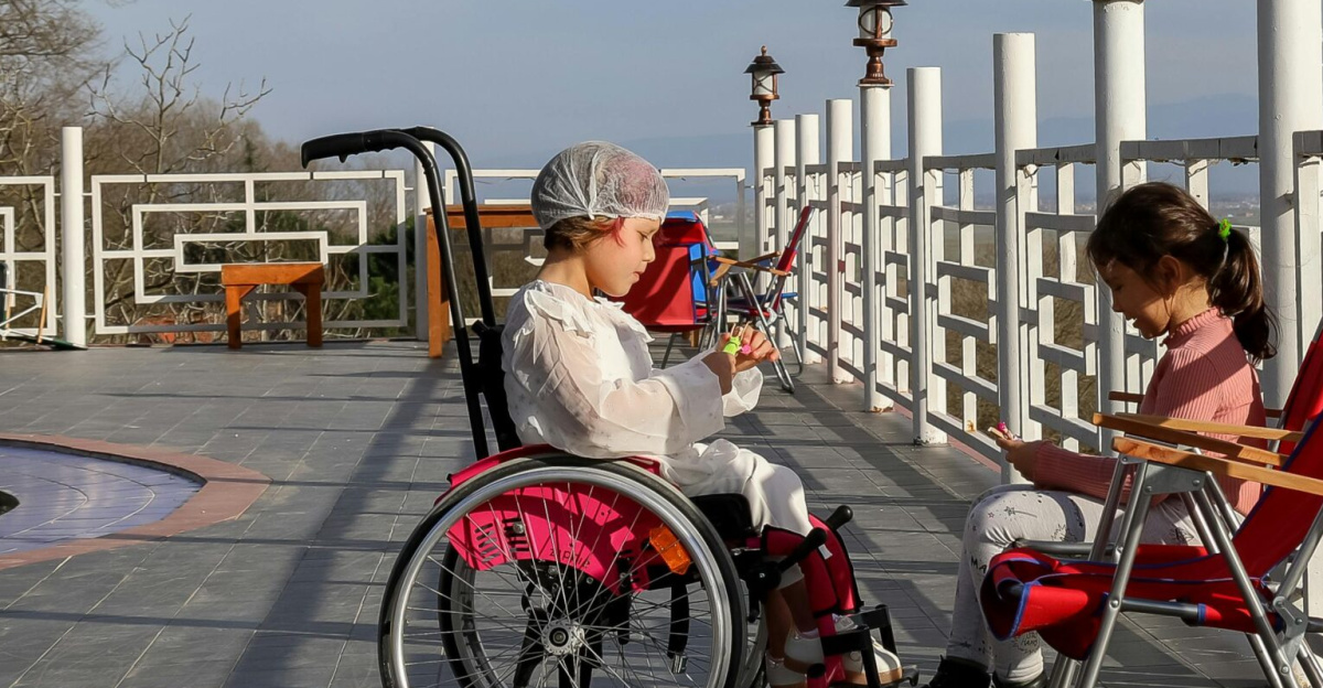Side view of girl in wheelchair speaking with friend in park under blue sky with clouds