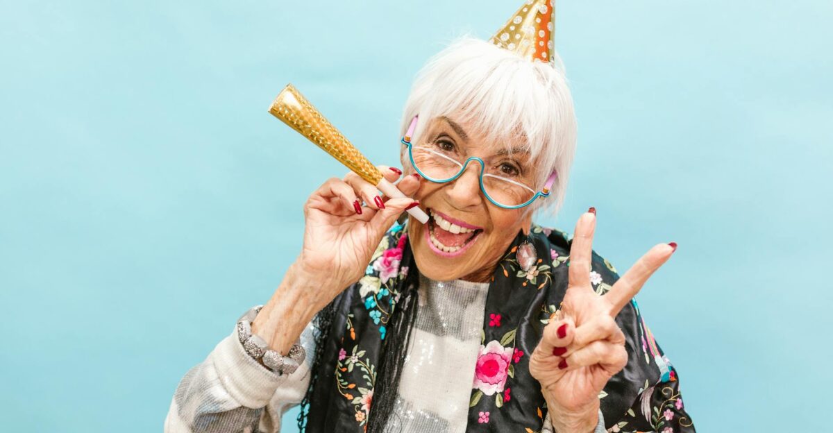 Senior woman in party hat joyfully celebrating making peace sign on a light blue background