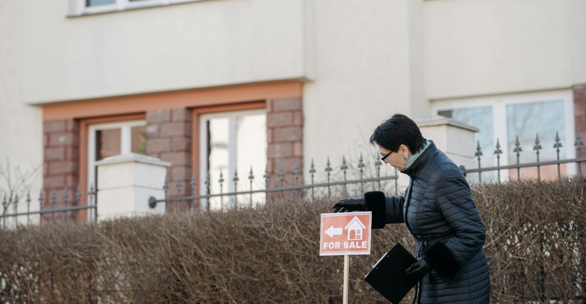 Real estate agent in a black coat placing a For Sale sign in front of a house