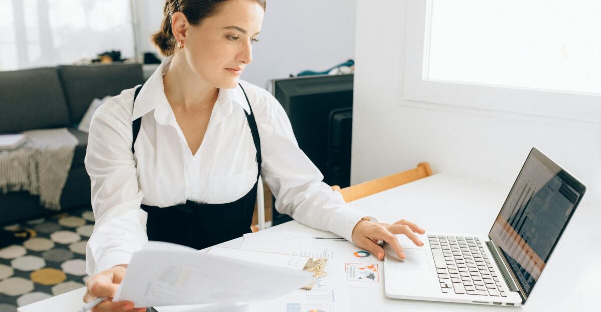 Woman at desk reviewing business reports on laptop holding papers in a modern office setting