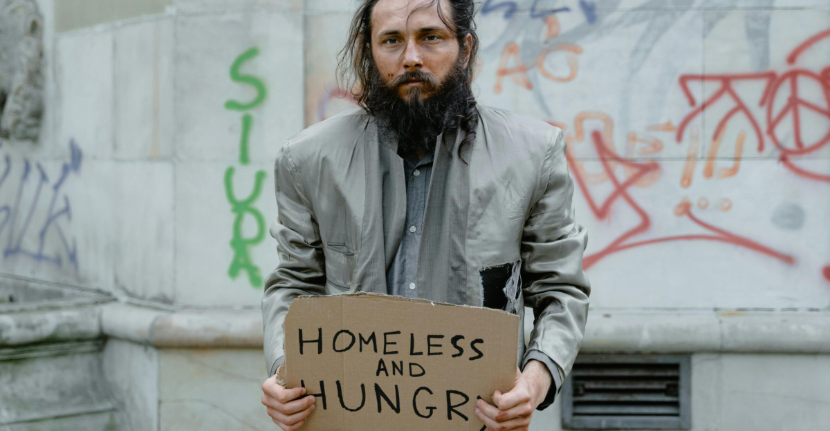 Homeless man standing on the street holding a sign, with graffiti in the background.