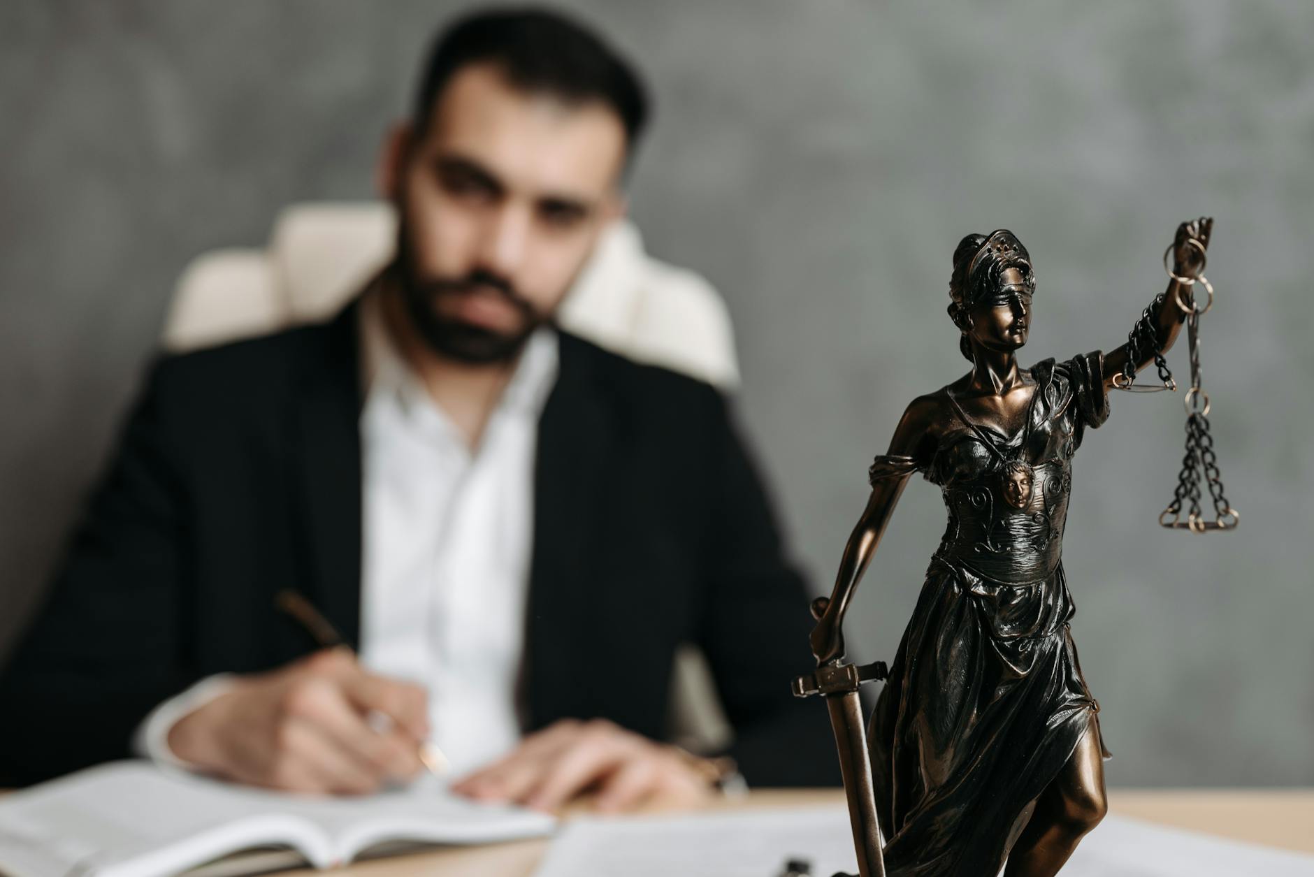 A lawyer in a suit writes at a desk with a Lady Justice statue in the foreground