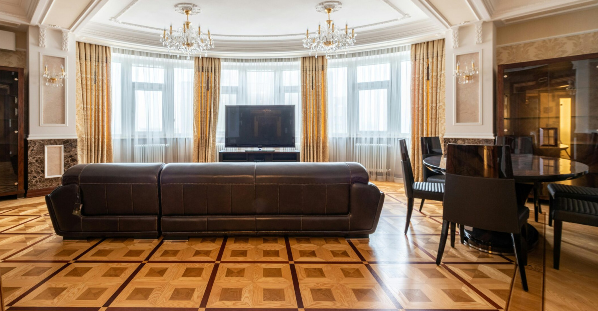 Spacious living room with a leather couch, large TV, and elegant decor, featuring chandeliers and parquet flooring.