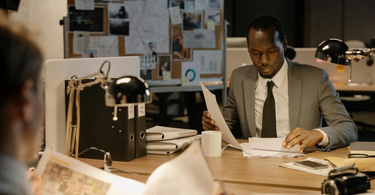 Detective in a suit examines papers at desk in a dimly-lit office suggesting investigation work