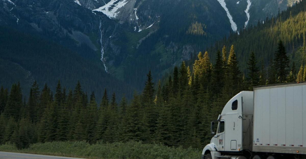 A semi-truck travels along a highway with snow-capped mountains in the background.