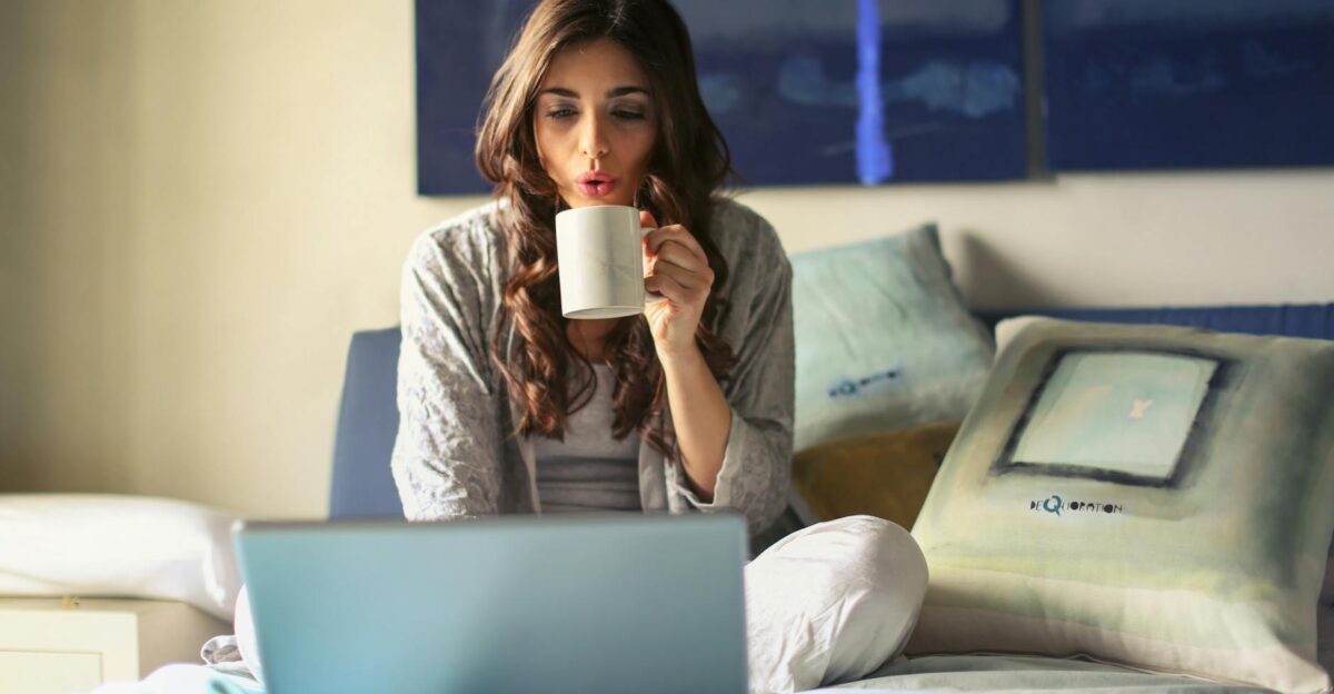 A woman enjoying coffee while working from home in a cozy bedroom setting with a laptop