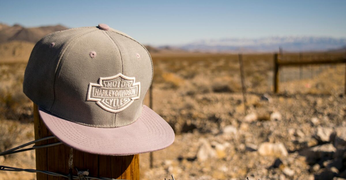A Harley Davidson cap sits on a fence post in an arid desert landscape under a bright blue sky