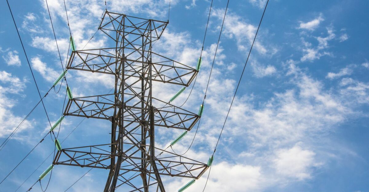 High voltage power line tower framed against a bright blue sky with clouds