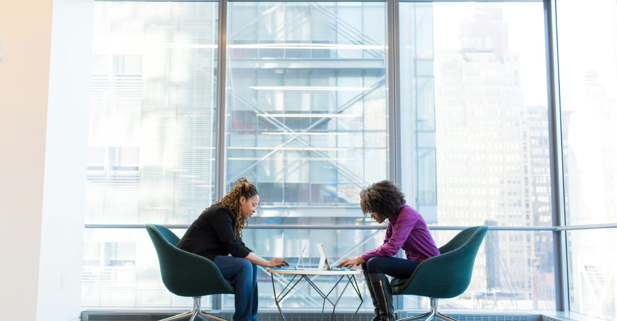 two women sits of padded chairs while using laptop computers