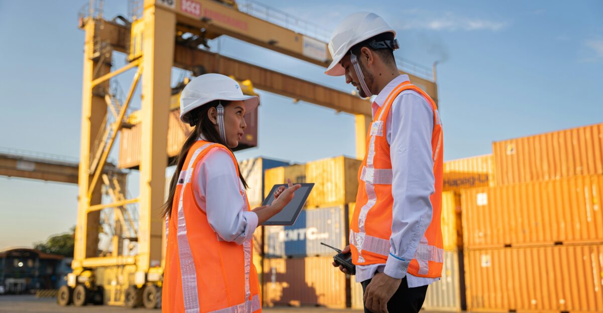Two workers in hard hats discuss plans near shipping containers