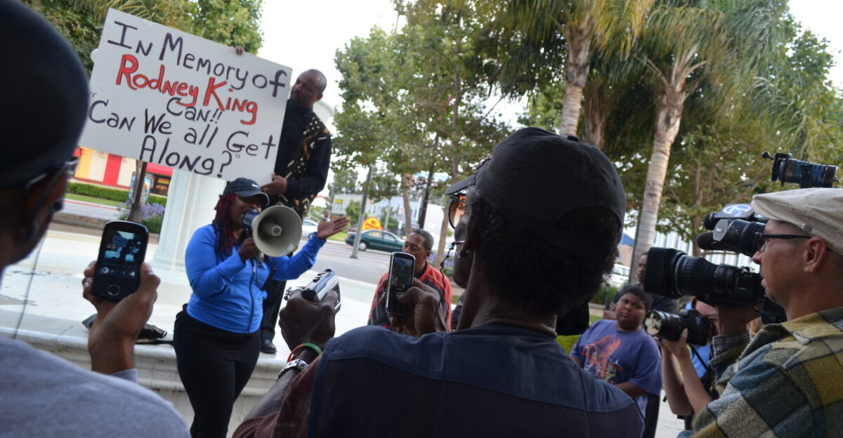 Activist Jasmine Cannock addresses a crowd of reporters and listeners Najee Ali supporting signage behind her
