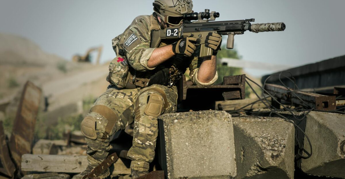 A soldier in full camouflage gear aiming a rifle while in a tactical stance outdoors